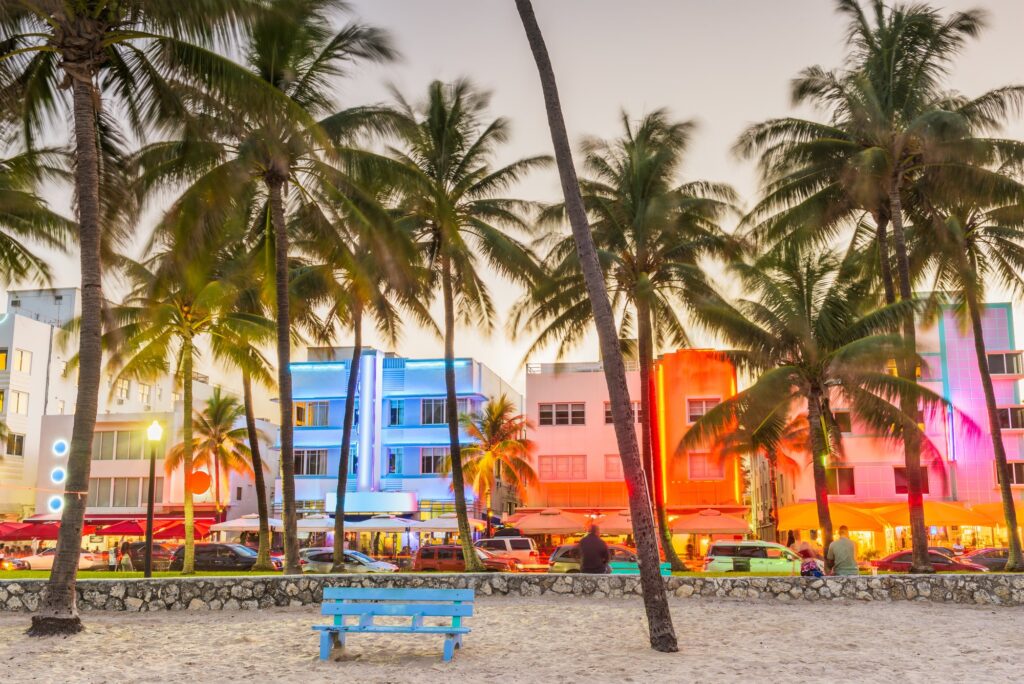 Neon-lit Art Deco hotels on Ocean Drive in Miami Beach at dusk, seen through palm trees from the sand